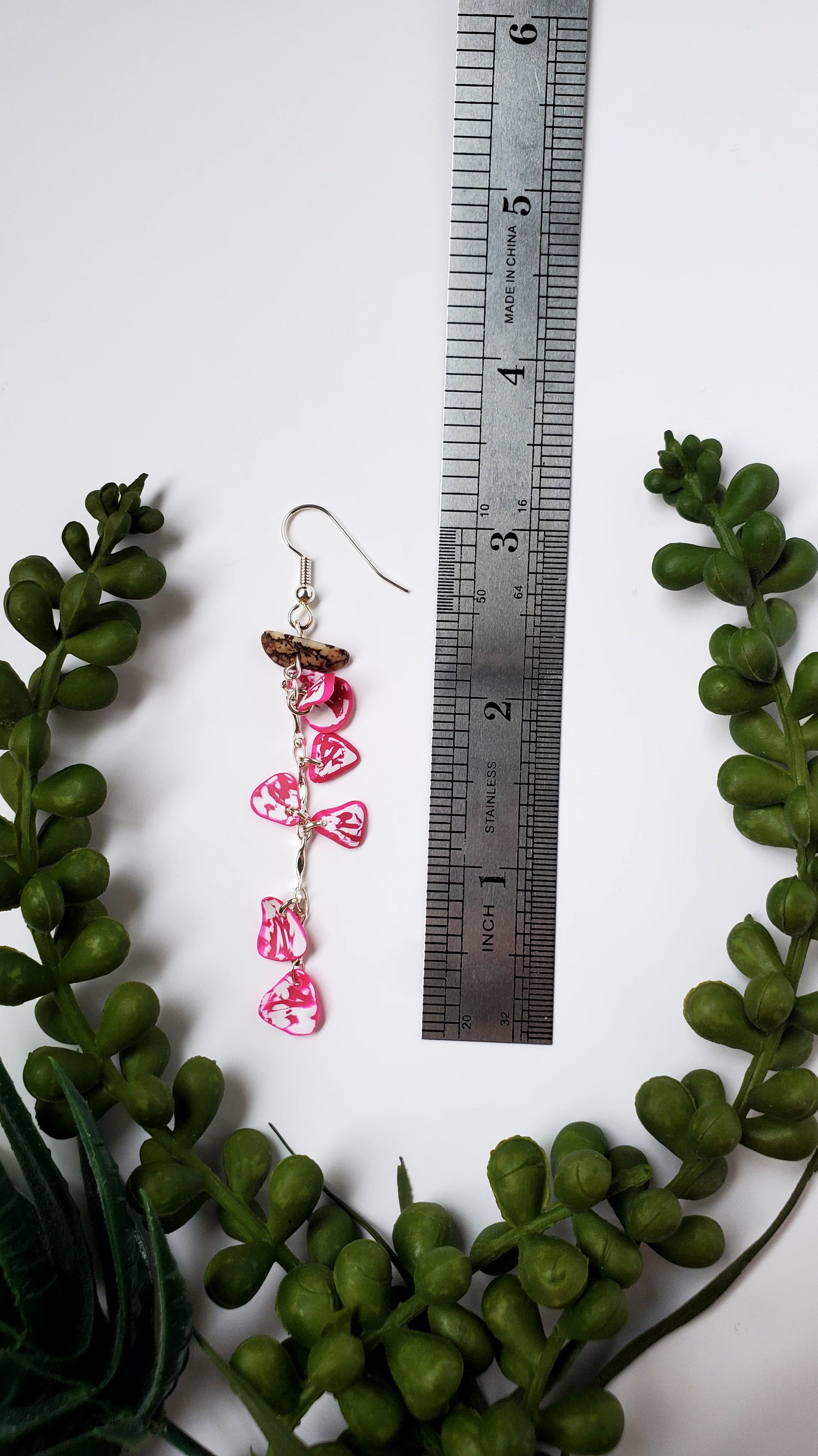 Pink butterfly earrings with a ruler for scale on a white background with green leaves.