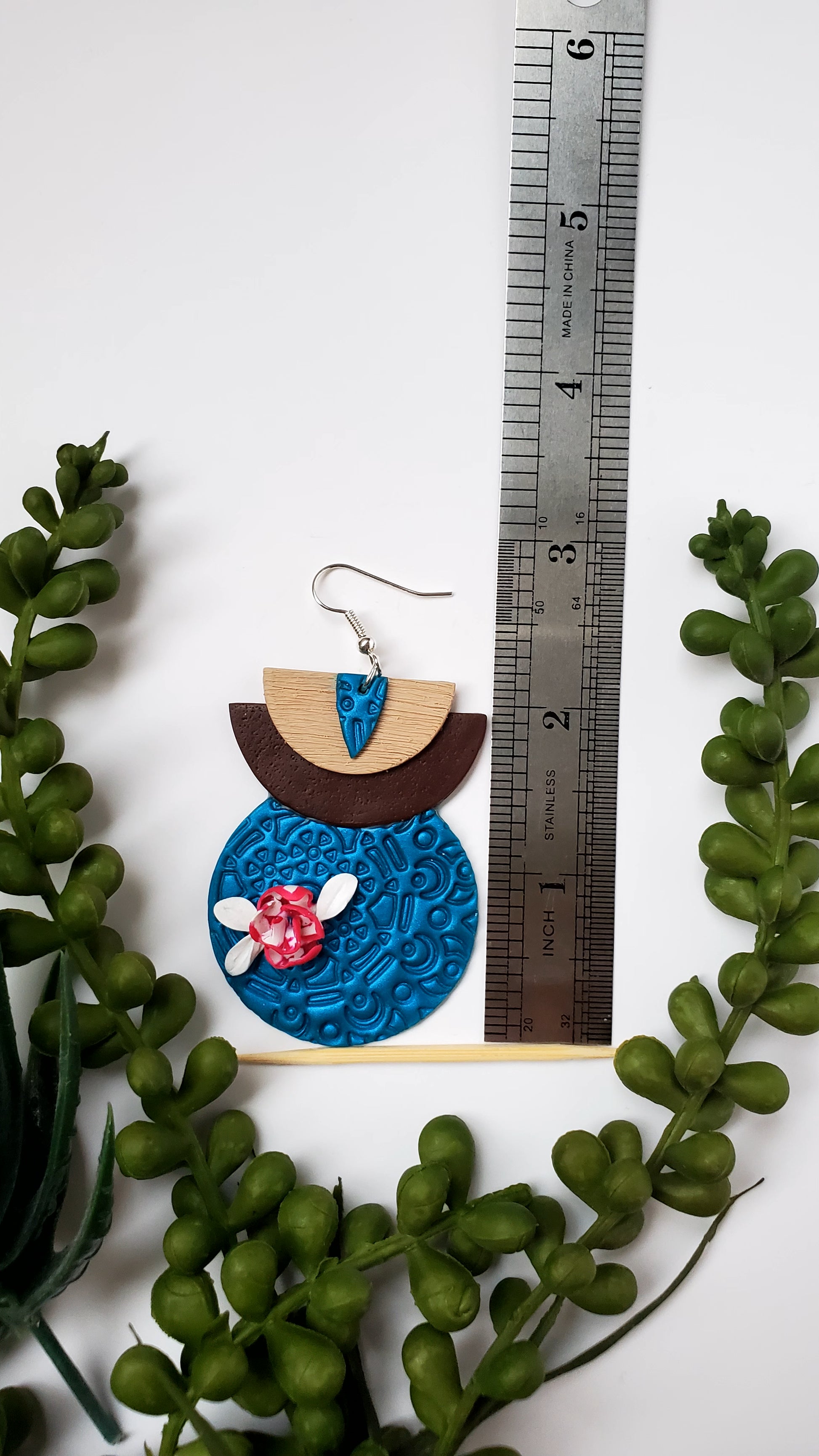 Earring with blue circular design and floral elements next to a ruler on a white background with green leaves.