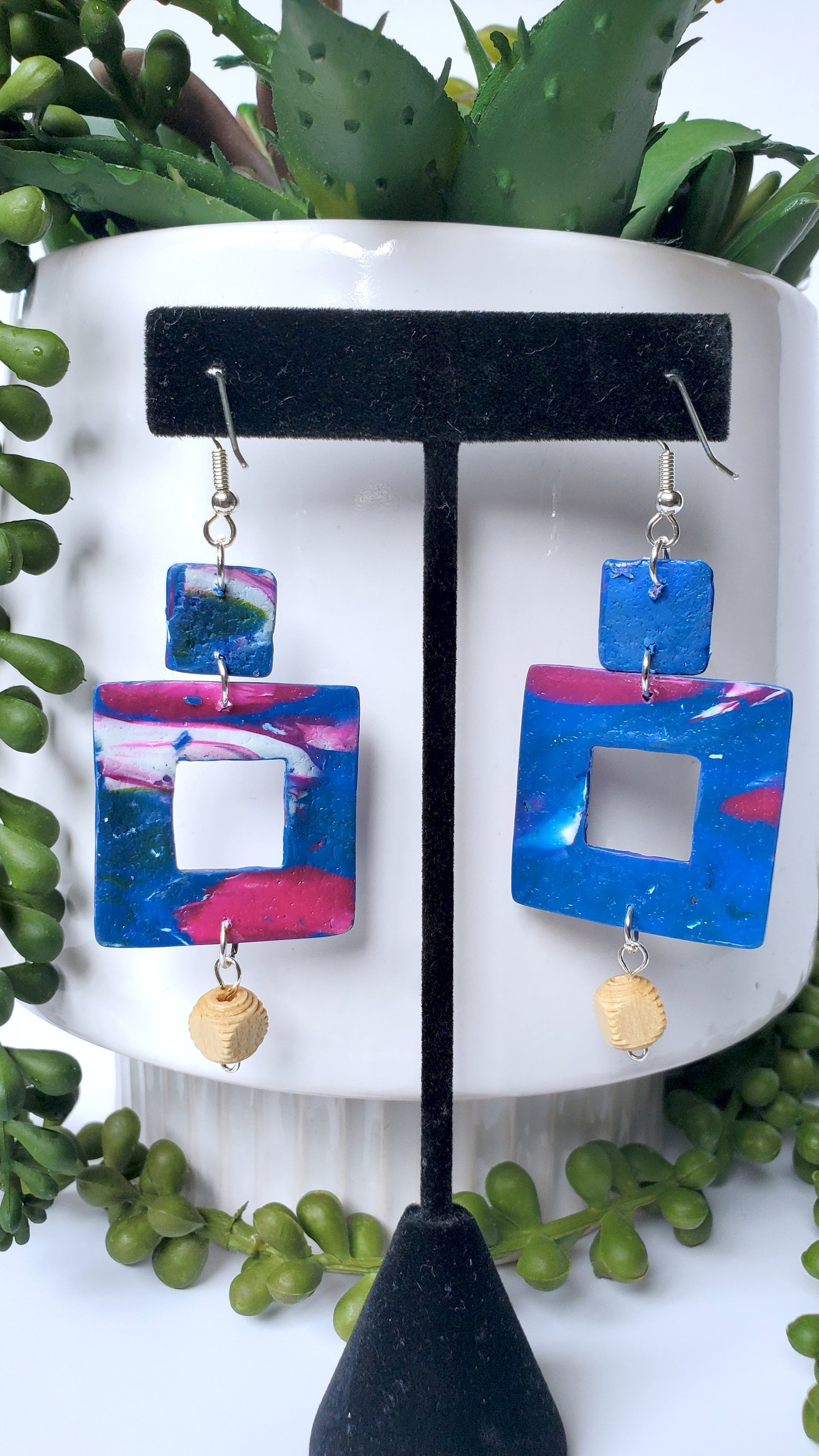 Colorful square earrings on a black stand with green foliage in the background