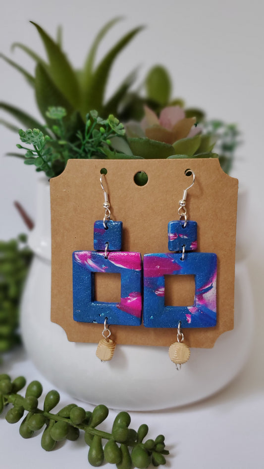 Colorful square-shaped earrings on a cardboard display card with green plants in the background.