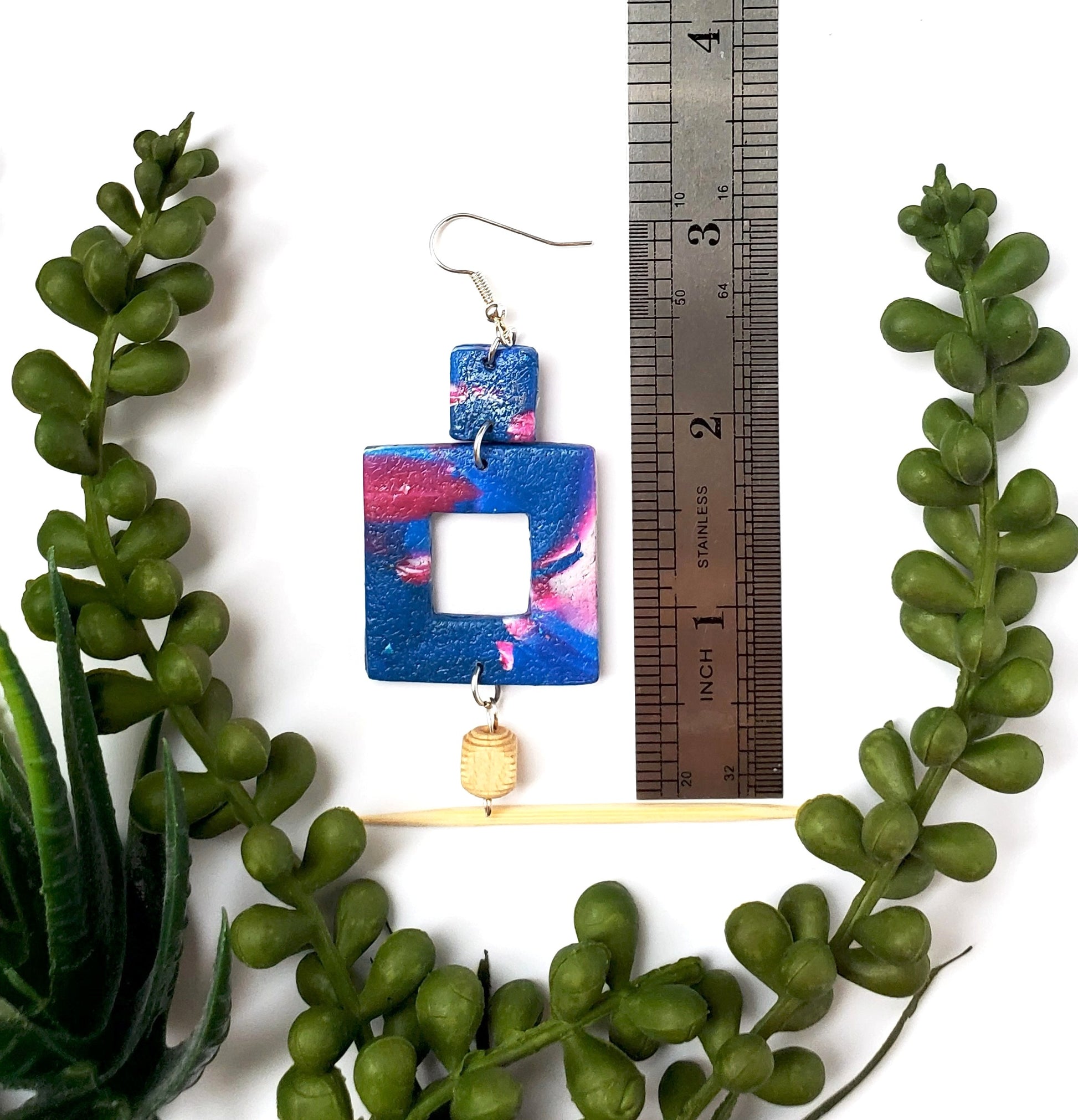 Colorful square-shaped earrings with a wood bead on a white background with green leaves and a ruler for scale.